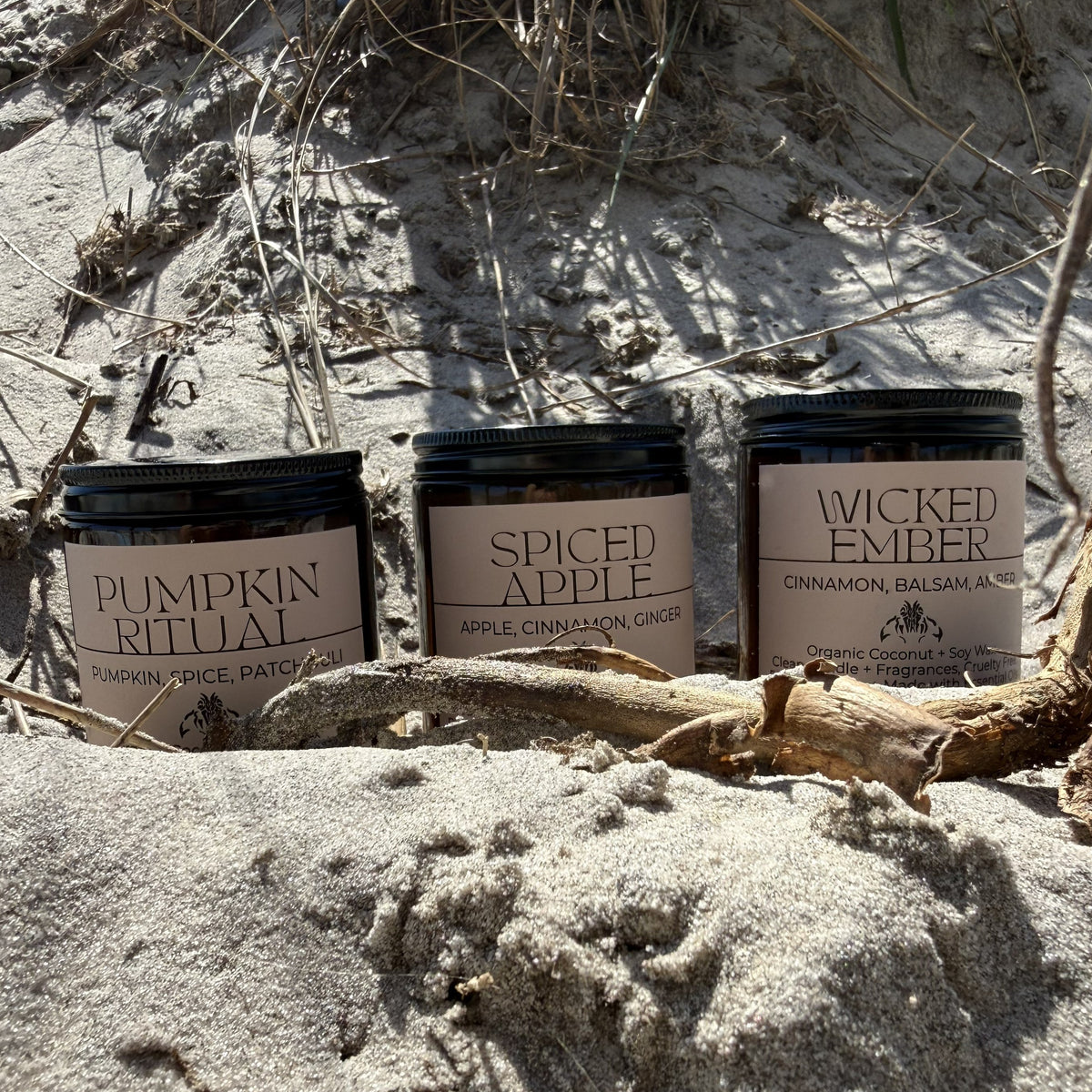 Three scented candles in amber jars for fall and halloween ambience, are arranged on sandy ground outdoors with beach grass and driftwood visible. The labels read Pumpkin Ritual, Spiced Apple, and Wicked Ember.